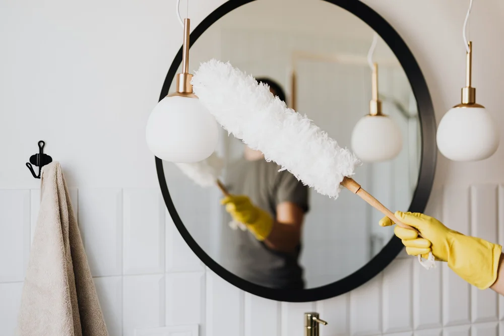 A duster carefully cleaning around a bathroom mirror and pendant lights